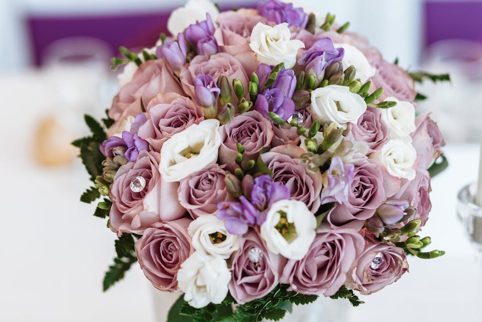 A close-up view of a floral bouquet featuring soft pink roses, white lisianthus, and purple freesias, arranged with green foliage. The flowers are fresh, with delicately textured petals and sturdy ste