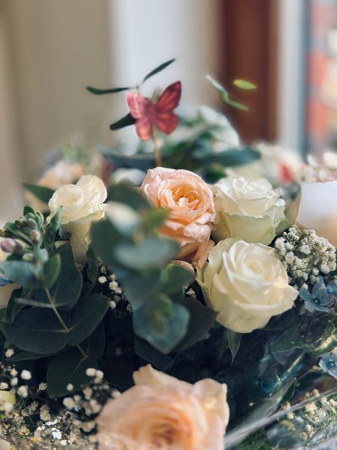 A close-up of a floral arrangement featuring cream and soft peach roses, surrounded by eucalyptus leaves and small white filler flowers, accented with a delicate pink butterfly decoration. The flowers
