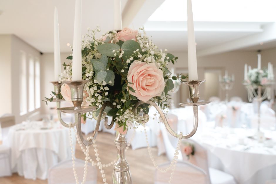 A close-up of an elegant floral arrangement featuring soft pink roses, white baby’s breath, and green eucalyptus leaves, arranged in a round bouquet style atop a silver candelabrum adorned with hang