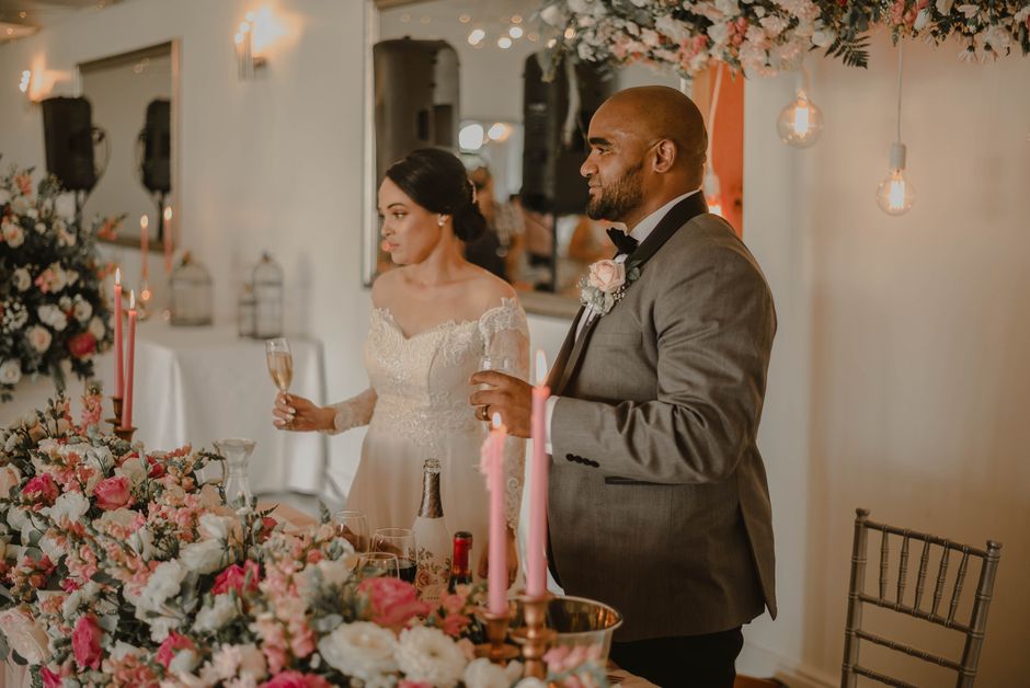 A bride and groom stand at a wedding reception table decorated with an elaborate floral arrangement featuring pink and white roses, peonies, and greenery. The bride, wearing a lace wedding gown with o