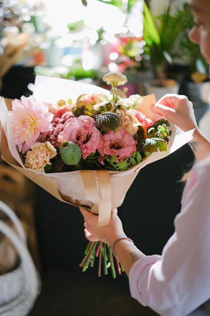A person holding a large floral bouquet featuring pink and cream peonies, roses, and chrysanthemums, accented with dark green foliage, wrapped in white paper with a beige ribbon. The arrangement is fr